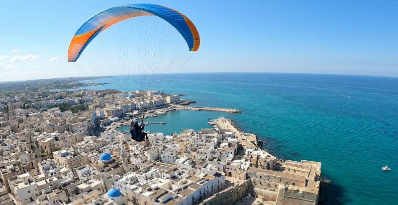 Pilota e passeggero in volo con il parapendio sopra le cupole azzurre e le spiagge di Monopoli